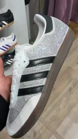 A person holds a white Adidas Samba sneaker with black stripes in their hand. The sneaker is adorned with sparkling rhinestones on the toe and heel. The sole is brown. The shoe is held in front of a store display with other shoes and clothing visible in the background. The person turns the shoe in their hand to show the details of the design.