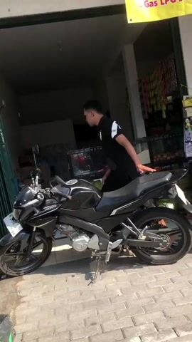 A young man wearing a black t-shirt and dark pants leans against a black motorcycle parked on a paved walkway. The motorcycle is positioned facing left in the frame, with its kickstand down. Behind the motorcycle, there is an open storefront with a green door. Inside the store, shelves are visible with various items stocked. A yellow banner hangs above the storefront, partially visible at the top of the frame. The man is looking downwards and to his right. The lighting suggests an outdoor, daytime setting.