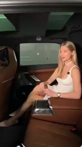 In a blue luxury car's tan leather backseat, a blonde woman in a white dress sits. The interior boasts screens behind the headrests and a sunroof. Two people stand outside the car. One is a woman with a long braid, holding a phone and taking pictures, while the other is a man wearing glasses, also holding a phone and looking at the car's interior. A third man stands behind him, partially visible. The scene takes place in what appears to be a garage, with fluorescent lights above. The woman in the car poses as photos are taken.