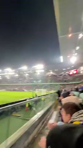 The video captures a scene from a packed stadium at night, likely during a sports event. Numerous spectators are seated in the stands, many are wearing casual attire like jackets and hoodies. The field, which is partially visible, is illuminated by stadium lights, and there's a scoreboard displaying the game's score.  A glass barrier separates the field from the stands. The stadium has a roof structure, enhancing the artificial lighting. The camera is held by someone in the crowd, providing a perspective from the spectator's point of view.