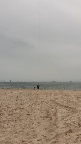 The video shows a person standing on a sandy beach, holding the lines of a kite. The kite is being raised into the air against a gray, overcast sky. The sea is visible in the background, with a few boats in the distance. The beach is sandy and has tire tracks in it. The person is standing still as the kite is raised.