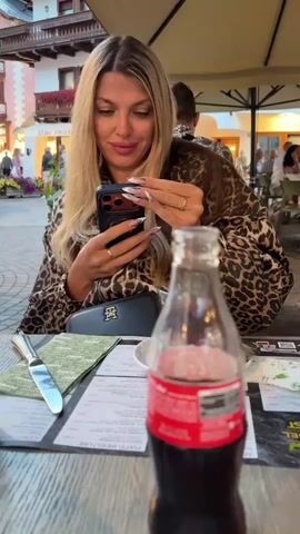 A woman with blonde hair and long fingernails sits at a table, looking at her phone. She is wearing a leopard-print top. In front of her is a glass, a menu, a knife, and a bottle of Coca-Cola. The background shows a European-style street with buildings and people walking around. A car is parked on the street. The scene appears to be an outdoor cafe or restaurant setting.