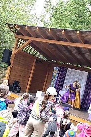 A woman in a purple and gold costume is dancing on a wooden stage, possibly performing for children. The stage is decorated with purple and white curtains. A group of children of various ages are watching the performance, some are dancing along, and adults are standing among the children. The stage is a small wooden structure with a roof. The environment appears to be outdoors, with trees visible in the background. A speaker and a laptop are visible on the left side of the stage. A white chair is on the stage. The word "WELCOME" can be partially seen on the stage backdrop.