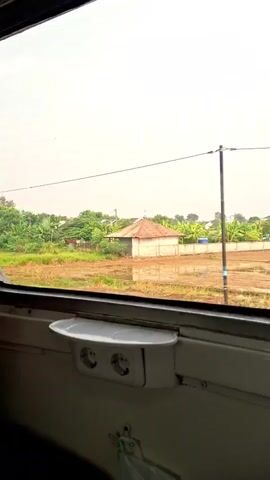 The view from a train window shows rice paddies and rural houses outside. The sky is overcast. Inside the train, there is a small shelf with two electrical outlets beneath the window. The train is moving, causing the scenery to pass by.