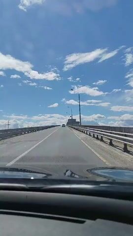 The video shows a first-person view from inside a car driving on a bridge. The road is paved with white lane markings and a metal guardrail lines the right side. The sky is a vibrant blue with scattered white clouds. Two cars are visible in the distance on the same road. A tall lamppost stands on the right side of the bridge, and other utility poles and structures can be seen in the background. The sun appears to be high in the sky, indicating daytime. The overall mood is peaceful and calm, suggesting a pleasant drive. There is no visible text in the video.