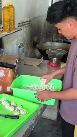 A young man with black hair, wearing a purple t-shirt and black pants with a yellow stripe, is in a kitchen preparing food. He is standing behind a bright green plastic container filled with white shredded ingredients, likely noodles or vegetables. He is holding a small portion of these shredded ingredients in his right hand and appears to be inspecting them. To his left, on another green plastic tray, are several round, white bundles tied with string, and a knife. In the background, there are two bottles of yellow cooking oil on a shelf, a large black wok on a stove, and other kitchen equipment. The environment appears to be a commercial kitchen with stainless steel surfaces and some areas showing signs of wear.