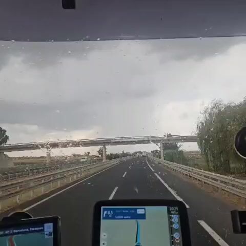 The video begins with a view from inside a vehicle, looking out through a rain-streaked windshield. The road ahead is a highway with multiple lanes, bordered by guardrails. An overpass is visible in the distance. In the foreground, two GPS devices are mounted on the dashboard, displaying maps and navigation information. The sky is overcast with dark clouds, suggesting rainy weather. The vehicle is in motion, as indicated by the blurred scenery and the onward progression of the road. A yellow truck is seen on the left side of the highway in the second frame. The video captures the experience of driving on a highway during a downpour, with rain distorting the view.