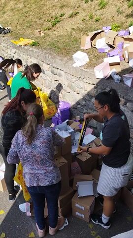 A group of people are gathered outdoors, appearing to be organizing or distributing items from boxes. The scene is set on an asphalt road next to a stone wall, with a grassy slope behind it. A silver van with its rear hatch open is parked on the left side of the frame, filled with boxes. Several individuals, including women and at least one man, are actively engaged in handling the boxes, some of which contain goods.

In the foreground, several people are focused on a collection of boxes and yellow plastic bags. One woman, wearing a patterned shirt, is reaching into a box. Another woman in a green shirt is looking down and seems to be interacting with a yellow bag. A woman with glasses in a dark t-shirt and striped shorts holds a yellow object with text that is partially visible.

In the mid-ground, a woman in a patterned dress and headscarf is interacting with the contents of the van. Other individuals are also seen near the van, assisting with the process. The ground is littered with some yellow triangular pieces and scattered pieces of cardboard.

In the background, a hilly landscape with vegetation and trees is visible, suggesting a rural or semi-rural setting. The weather appears sunny, indicated by the shadows cast on the ground. The overall atmosphere seems to be one of organized activity, possibly a donation or aid distribution event. The text on one of the boxes is partially visible and appears to be "Gala" and "bananas". The time of day appears to be daytime, likely late morning or afternoon, given the sunlight.