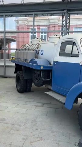 The video shows a vintage blue and white truck with a large tank on the back. The truck is parked on a paved surface, possibly a museum exhibit or outdoor display. The background shows a building with arched windows and glass structures. The camera pans from the rear of the truck to the front, focusing on the details of the vehicle, such as its tires, headlights, and the writing on the tank, which appears to be in Cyrillic letters and says "МОЛОКО" (milk). There are no people visible in the video.