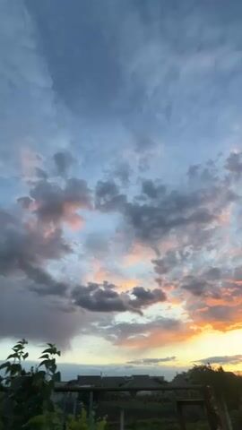The video captures a picturesque sky during sunset. The sky is predominantly blue but filled with a mix of gray, orange, and pink clouds. In the lower portion of the frame, the silhouette of a residential area is visible, featuring houses and greenery. The lighting suggests it's either early evening or dawn. The clouds exhibit soft hues, indicating the sun is either setting or rising.