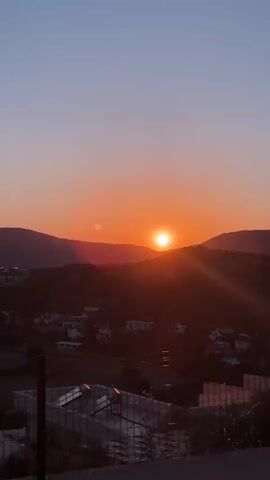 The video captures a serene sunset over a mountainous landscape near Kaş, Turkey. The sun, a brilliant orange orb, gradually descends behind the distant mountains, casting warm golden and orange hues across the sky. Below the mountains, a small town or village with white buildings is nestled in the valley, partially visible amidst the green, bushy terrain. A prominent red lens flare appears in the lower-left portion of the frame for most of the video. As the sun continues to set, the camera appears to be in motion, as evidenced by a tree branch quickly entering the frame from the left and blurring as it passes, momentarily obscuring the view of the sunset before the video ends. The sky transitions from a deep blue at the top to a vibrant orange and yellow near the horizon, indicative of a clear evening. No people or visible text are present in the footage.