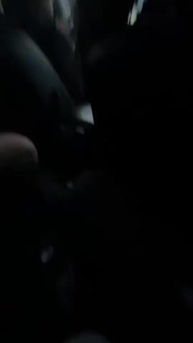 The video shows the interior of a car in motion. There are at least three people inside, their faces are obscured but their head and shoulder outlines are visible. The interior is dark, the seats are black. One person in the back is holding and looking at a smartphone. There's music playing. The outside environment is visible through the car windows and appears to be mostly overcast. The car seems to be moving on a road outside a populated area.