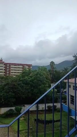 The video shows a climb up a blue metal staircase. The staircase has vertical railings. At the top of the staircase, there is a balcony with a blue railing. The balcony is connected to a building with a red tile roof. A large green tree is visible next to the building. In the background, there are green trees and a mountain range under a cloudy sky. The day appears overcast. There is a window open next to the balcony. The building’s exterior is painted white.
