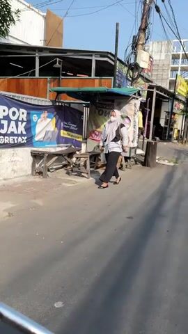 A woman wearing a light-colored hijab, a grey t-shirt, and black pants walks on a paved road. She is carrying a grey bag over her shoulder. She is walking away from the camera. To her left, there is a food cart with a blue and white banner that has text on it and an image of a man. To her right, there is a wooden pole and an alleyway. In the background, there are buildings and overhead wires. The sky is blue and it appears to be daytime.
