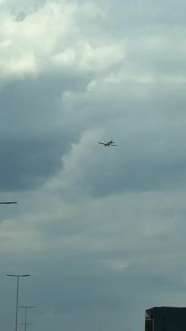 A small yellow airplane flies through a cloudy sky over a highway. Several cars drive on the highway. Streetlights line the side of the road. Mountains are visible in the distance.