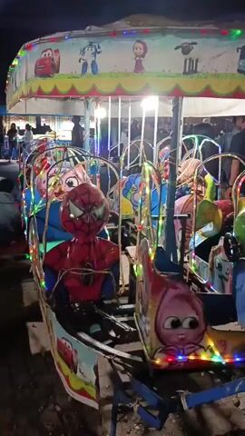 A short video clip shows a children's amusement ride, likely a carousel or spinning train, at night. The ride is adorned with colorful lights and features various cartoon characters on its seats, including Spider-Man, a pink cat, and other animated figures. People are visible in the background, suggesting a public event or fairground. The ride appears to be in motion, with the seats swinging and rotating. The environment is dark, with the artificial lights of the ride and surrounding areas illuminating the scene. The ride seems to be made of metal and plastic, with a colorful canopy overhead decorated with more cartoon imagery.