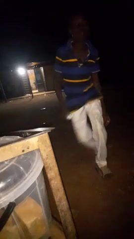A person is captured in a low-light video, appearing to be outdoors at night. They are wearing a blue and yellow striped short-sleeved shirt and white pants. The person is in motion, with their body blurred due to movement. They are holding a light-colored object in their right hand. In the foreground, a portion of a large clear plastic container with a wooden frame is visible. The background is dark with some structures and lights suggesting a residential or outdoor setting. The person's facial features are indistinct due to the lighting and blur.