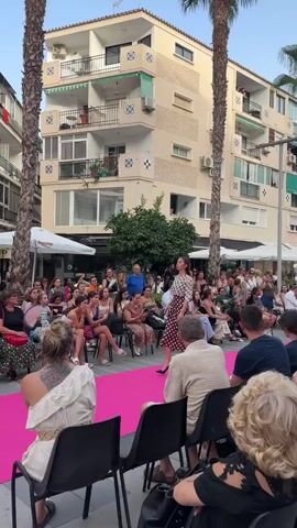 A fashion show is taking place outdoors on a street. A woman wearing a polka dot dress and high heels walks down a bright pink runway. She is surrounded by an audience sitting in chairs, some of whom are holding fans. Palm trees are visible on either side of the runway, and buildings with balconies line the street in the background. The setting appears to be a town square or pedestrian street.