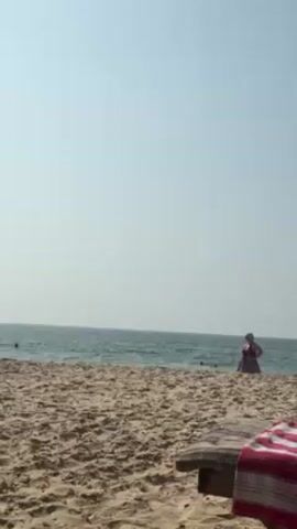 On a sunny beach, a man is sitting on a black motorcycle that is on the sand near the water's edge. He is shirtless and wearing a cap.  A woman in a red dress is standing near the water. There are some people in the water swimming.  A striped towel is on a bench or mat in the foreground.  The sky is clear.