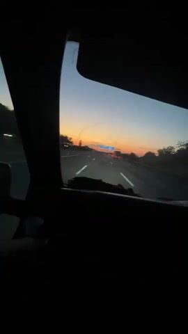 The video captures a view from inside a vehicle looking forward through the windshield at dusk. The sky displays a gradient of colors from deep blue overhead to orange and yellow near the horizon, indicating sunset. A highway stretches ahead, with multiple lanes of traffic visible. Cars and trucks can be seen in the distance. A large blue overhead road sign with white text is illuminated. The foreground shows the dark interior of the vehicle, including the dashboard with some blue LED lighting. The perspective suggests the vehicle is in motion, traveling on the highway.