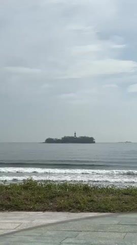 A video shows a wide shot of a calm ocean with a small, tree-covered island in the distance. A lighthouse stands on the island. The water is a greyish-blue, and gentle waves are breaking on the shore in the foreground. Green grass and a paved path are visible in the bottom portion of the frame. The sky is overcast with light grey clouds. No people or animals are visible in the video. The camera is stationary.