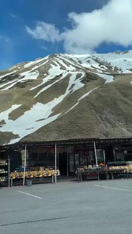 The video shows a mountain range on a sunny day, partially covered in snow. In the foreground, a roadside stand with fruit and souvenirs is visible. A blue portable toilet is next to it. The camera moves slightly to the right, revealing a red and white road sign and a parked white car. The focus then shifts to the mountain, emphasizing the patches of snow interspersed with brown vegetation. The sky is blue with scattered white clouds.