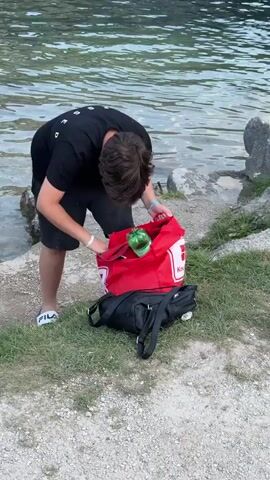 A young person with brown hair, wearing a black t-shirt and black shorts, is standing on a rocky shore next to a body of water. They are holding a large red tote bag with white trim and a white logo, and they appear to be putting something into or taking something out of it. A black bag is lying on the grass near the person's feet. The water in the background is calm and appears to be a lake or ocean. The ground is a mix of gravel and grass. The person is wearing white slides with a logo. They are also wearing a blue wristband. They look towards the camera and speak.