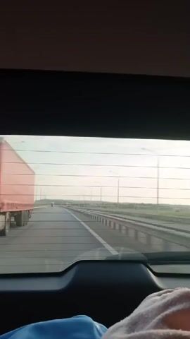 A video taken from inside a vehicle, looking out the rear window. The road ahead is a multi-lane highway. A white semi-truck with a red trailer is traveling in the left lane. In the distance, there are streetlights and a green signpost. The sky is bright, suggesting daytime. A person's legs and foot, wearing a pink blanket and blue clothing, are visible in the lower right corner of the frame, indicating the perspective is from a passenger.