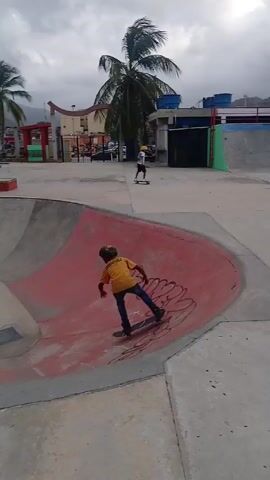 A young child on a skateboard is riding in a skatepark, moving from the bowl area toward some low rectangular ledges. The child wears a yellow shirt and helmet. In the background are buildings, palm trees, and people on benches. The sky is overcast.