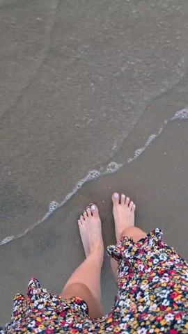 The video shows a first-person perspective of someone walking into the ocean. Their feet and lower legs are visible, wearing a floral-print dress. The water is shallow and clear, with small waves and foam washing over their feet. The sand on the seabed is also visible. The camera movement follows the person's steps into the water.