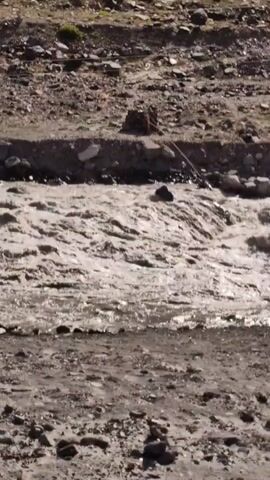 A muddy river flows through a rocky, barren landscape. The water is turbulent and churns with whitewater, indicating a strong current. The riverbanks are composed of loose soil and rocks, with some vegetation sparsely scattered. A rope is visible, stretched across the river from the bank. A small, dark object, possibly a large rock or debris, is submerged in the water, creating a visible disturbance in the current. The overall impression is one of a wild and untamed natural environment.
