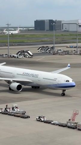 A China Airlines airplane is parked on an airport tarmac. Multiple luggage carts are being towed by a tug towards the plane. In the background, other airplanes, stairs, buildings, and control towers are visible. The sky is cloudy.