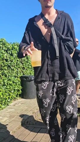 A man dances on a wooden deck outdoors in bright sunlight. He wears a black hat, sunglasses, black, patterned pants, an unbuttoned black shirt, a black bag slung across his chest, and a silver necklace. He is holding a yellow beverage in a clear cup. He moves his hands in sync with the music, keeping his eyes forward. Behind him, a green hedge provides a backdrop. A black trash can sits to his left. A few other people, including a man in shorts, are visible further in the background. The sky is bright blue.