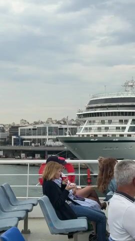The video shows a shot from the deck of a boat, possibly a ferry, with passengers seated on plastic chairs. In the background, a large cruise ship named "Viking Star" is docked. A woman in a black jacket and jeans sits in a blue chair, eating from a cup and looking at her phone. A man in a white shirt and blue shorts walks by, talking on his phone. To the right of the woman, another passenger with curly hair sits in a similar chair. In the foreground, part of the head of another passenger is visible. A red life preserver is mounted on the railing of the boat. The sky is overcast. Buildings are visible in the distance behind the cruise ship.