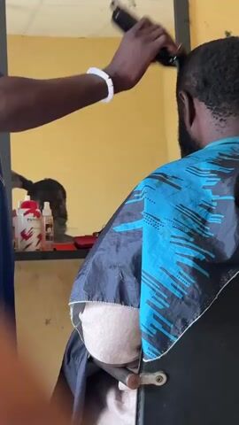 A barber cuts a man's hair from behind. The man is seated and wearing a blue and black cape. The barber is using clippers and is partially visible in the frame. The background appears to be a room with yellow walls. There are some bottles and a mirror on a shelf in the background.