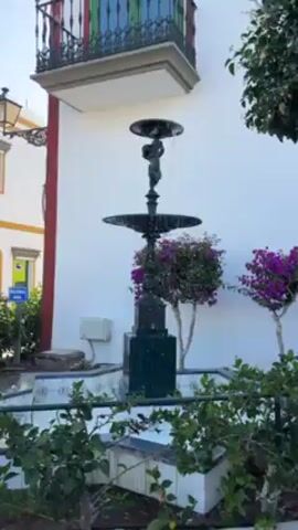 The video shows a fountain with a statue of a child on top, surrounded by greenery and set against a white wall. The fountain has multiple tiers from which water flows. The base of the fountain is surrounded by a tiled area and various plants. The setting appears to be an outdoor, possibly urban, environment.