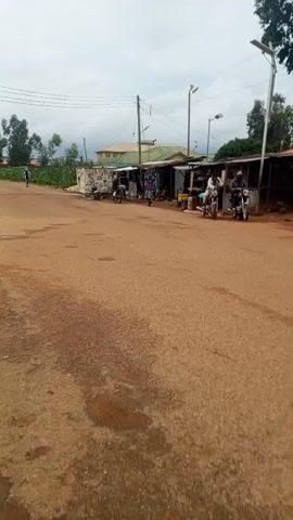 The video pans across a dirt road with some scattered gravel and puddles. On the right side of the road, there are several open-air market stalls or shops constructed with corrugated metal roofs and wooden supports. Several motorcycles are parked near these stalls, and a few people are visible standing or interacting around them. On the left side of the road, there are some buildings and what appears to be a concrete drainage ditch. In the background, there are trees, more buildings, and utility poles with overhead wires. The sky is overcast, suggesting a cloudy day. There are people walking and riding motorcycles on the road in the distance as well.