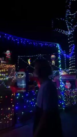 At night, a house is heavily decorated with Christmas lights.  A young person stands in front of a fence, viewing the display.  Inflatables of Santa Claus wearing sunglasses sit on the front lawn, with more Santa inflatables on the roof.  The house is covered in blue, white, yellow, and red lights.  A tree is wrapped in colored lights.  The camera pans across the house and the yard.  There is a Christmas sign in the background.