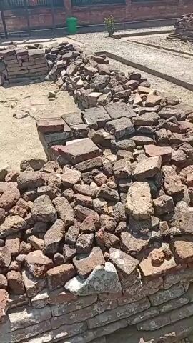 The video shows an outdoor archaeological site with piles of bricks and rocks scattered around. In the background, there is a building with a tiled roof. Several people are present, some standing and others sitting near a motorcycle. One person is holding up a phone, possibly taking a picture or video. The ground is dusty and appears to be a mix of dirt and sand. The overall impression is one of an ongoing excavation or preservation effort at an ancient ruin.