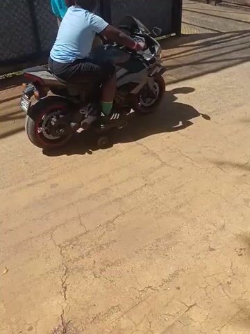 A man is seated on a parked motorcycle, which is angled slightly to the left in the frame. A second man in a blue shirt stands beside the motorcycle, next to a black metal gate. The motorcycle is primarily gray with red accents on the wheels. The ground is a cracked, tan-colored concrete surface, with tree shadows cast across it. A low wooden fence is visible in the background. The camera's perspective is from above, looking down at an angle, giving a slightly disorienting view.