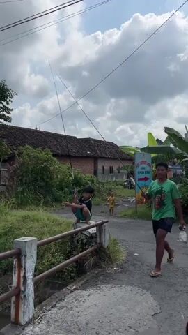 A wide shot shows a rural scene with several children fishing. Two boys are in the foreground, one in a green t-shirt and the other in a blue t-shirt, both holding fishing rods and looking towards the water. In the background, another child in a red shirt is also fishing, and a motorbike is parked near a building. The environment includes a bridge railing, a canal, lush green grass, trees, power lines, and a cloudy sky.