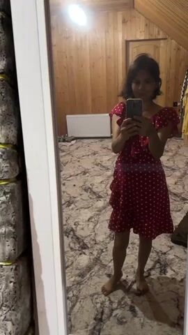 A woman in a red and white polka dot dress stands in front of a mirror, taking a selfie. The room has wooden walls and a patterned floor. A radiator is visible on the wall behind her. She is barefoot and has short dark hair. She adjusts her hair as she takes the photo.