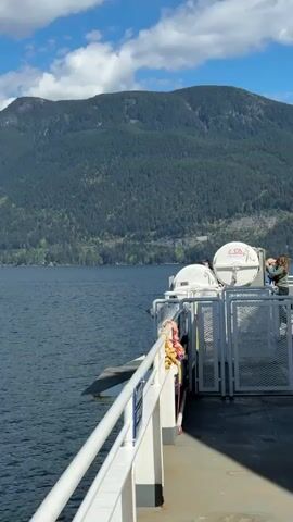 The video depicts a ferry journey on a clear, sunny day. The camera pans across the water, revealing mountainous terrain in the distance, partially covered with trees and some snow at their peaks. The water is a deep blue, slightly choppy. The foreground shows the side of the ferry, including a railing and a white structure, possibly a life raft container. The sky above is a vibrant blue with scattered white clouds. The overall impression is that of a serene and scenic ferry ride.