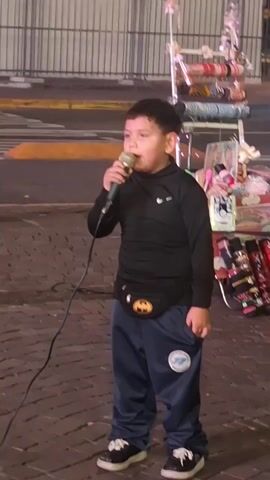 The video shows a young boy singing into a microphone on a brick-paved street. He wears a black long-sleeved shirt, blue pants with a patch, and black and white shoes. A Batman fanny pack is around his waist. He stands in front of a rolling cart displaying various items. The background includes a street with cars, a tall monument, and other pedestrians. The boy sings with his hand occasionally in his pocket. The camera moves slightly, showing more of the surroundings.