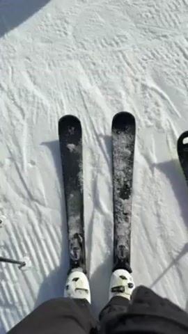 The video shows a group of people skiing on a sunny day. The first shot is of the skis, then the camera tilts up to show other skiers on the hill and the view of snowy mountains in the background.  There are at least five skiers visible, all wearing winter clothing, helmets, and goggles. They are standing on a well-groomed ski slope covered in snow. The landscape behind them shows a vast mountain range under a bright, sunny sky.