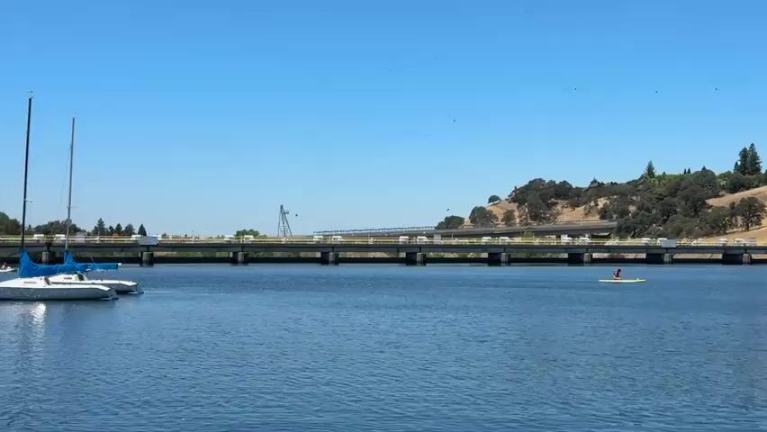 A wide shot shows a calm, blue lake on a sunny day with a clear sky. In the foreground, on the left side of the lake, two sailboats are docked. One is white with blue sails, and the other is partially visible behind it. In the middle ground, a long, dark bridge stretches across the water. Cars can be seen driving on the bridge. On the right side of the lake, a person is paddling a yellow kayak. The background features a green, hilly landscape with scattered trees. The overall scene is serene and depicts a recreational area.