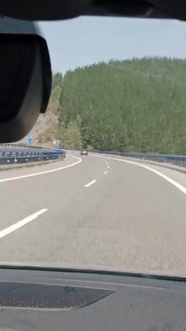 The video is a first-person perspective from inside a vehicle driving on a highway. The road is paved with asphalt and has white lane markings. There are guardrails on either side of the road, painted blue and silver. A single car is visible ahead on the highway. On the right side of the highway, there is a forested hillside with many pine trees. On the left side, there is a barren, rocky hillside with some vegetation. The sky is clear and blue. There is a black object, possibly a dashcam or mirror, attached to the windshield or dashboard, partially visible in the upper left corner of the frame. The interior of the car, including the dashboard and part of the windshield, is visible in the foreground.