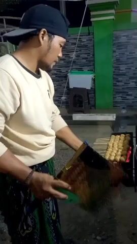 A man wearing a black baseball cap and a light-colored long-sleeved shirt is standing next to a grill and fanning it with a flat object. He is also holding a torch in his left hand. The grill contains several skewers of food, which appear to be meatballs. The food is being cooked over hot coals. The man is standing on a dirt or gravel surface, and there is a wall with green and black tiles behind him. The scene appears to be outdoors, possibly at night, as indicated by the lighting.