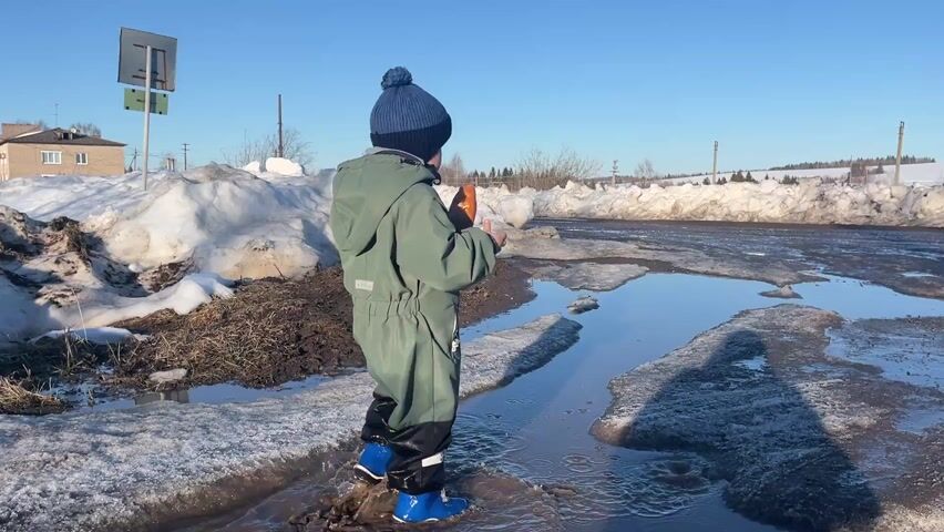 A young child in a green jumpsuit, black rubber leggings, and blue boots stands in a large puddle on a sunny day. The child wears a blue knitted hat with a pompom. Snow banks line the sides of the muddy road and the sky is clear and blue. The child takes a small orange object from a pocket, holds it up to their mouth, and then puts it back. The child is looking towards the snowy landscape ahead. A road sign and a building are visible in the background.