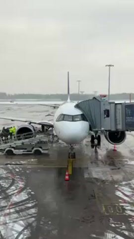 An airplane stands at an airport gate. The day appears to be overcast and the ground is wet with traces of snow. A jet bridge is connected to the aircraft, and a baggage conveyor belt is positioned near the plane's engine.  A person wearing a high-visibility jacket is visible near the baggage conveyor. The airport tarmac shows streaks of water and residual snow. The jet bridge has an advertisement on its side.  The overall scene depicts ground operations occurring around a parked airplane at an airport under wintry conditions.